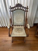 Front view of Eastlake style folding chair with visible tapestry upholstery on backrest and seat, carved wooden frame and padded armrests.