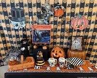 Wide shot showing an assortment of Halloween decorations arranged on a black and orange supernatural-themed cloth featuring stars and moons. Items include skull figurines, bats, witches, pumpkin faces, ghost signs, skull mugs, and spiders.