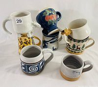 Nine assorted hand-thrown coffee mugs with varied artistic glazes and designs in different shapes and sizes, stacked in a cluster.