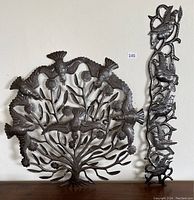 Two metal wall hangings displayed against a wall. Left is a circular tree of life design with birds; right is a vertical birds on branch design.