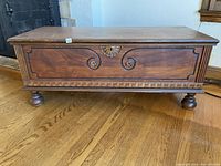 Front view of wooden cedar chest showing the carved front panel with scroll and shell designs, rounded bun feet, and wear marks on the top surface.