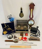 Full lot showing wooden barometer, brass weight scale, inkwell desk set, Polaroid camera, office supplies, retractable tools, and teachers bell.