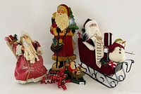 Lot showing three Santa themed decorations together on a white backdrop: a jolly fabric Santa table topper, a wooden Santa tree topper with a staff, and a wooden Santa figurine with a full beard, next to a Santa in sleigh with snowman figure.
