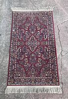 Full view of one of the Karastan Red Sarouk rugs on concrete showing detailed floral and geometric pattern in red, navy, cream, and beige colors with fringed ends.