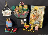Overview of Easter decor lot including decoupage eggs in basket with green Easter grass, the blue egg stroller with bunny and chick figures, wooden duck figurine, woven baskets, and bunny egg-cups on display.