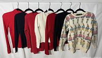 Five sweaters hanging on hangers on a white backdrop, showing color and style variety