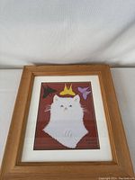Photo of framed Maud Lewis print showing white cat with butterflies, wooden frame, and off-white matting.