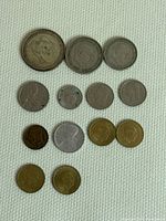 Photo showing 15 various European coins laid out on white fabric, different sizes and metals, with visible portraits, numerical denominations, and emblems.