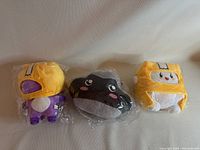 Front view of three unopened plush stuffed toys in plastic packaging, two yellow and one black in color.