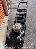 Full view of green and metallic lawn mower with gas engine and foldable handlebar, parked outdoors outside a facility.