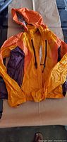 Full view of the orange and purple women's windbreaker jacket lying flat, showing hood, sleeves, and color block design.