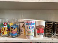 Five 1980s McDonald's glasses, 1986 Miami Vice 7-11 cup, 1985 Jetsons/Orange Julius cup, and other promotional glasses grouped on a shelf