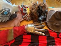Photo showing the Freddy Krueger costume sweater, claw glove, and four masks including a polar bear or wolf mask, a colorful Luchador mask, a werewolf mask, and a brown fedora hat