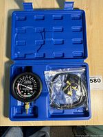 Blue plastic carrying case opened to show black round vacuum gauge, clear dial with markings, and sealed plastic bag containing hoses and brass connectors.