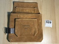 Full view of the brown leather tool bag with various compartments and pockets visible, laid flat on a wooden surface.