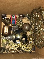Box filled with assorted brass decorative items including mask, bells, and figurines, viewed from above inside a cardboard box.