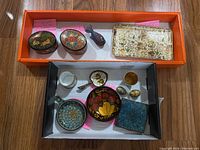 Overview of trinket boxes, enamelware cups, small painted spoon, and floral patterned metal tray arranged inside two boxes