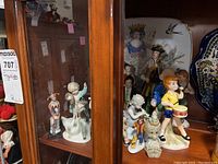 Photo shows multiple ceramic figurines of various styles and sizes displayed inside a cabinet shelf, with a decorative plate in the background.
