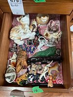 View of drawer with a large orange and white ceramic cat figurine lying down, surrounded by multiple smaller cat figurines made of ceramic, fabric, and painted metal, along with a green cat figurine on a wooden base.