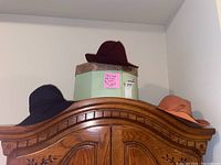 Three wool felt hats displayed on an antique wooden wardrobe: maroon, navy blue, peach colors.
