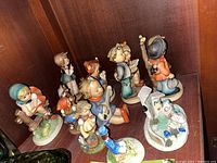 Group photo showing nine ceramic figurines displayed on a wooden shelf, depicting children in various poses and traditional clothing.