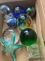 Full lot of 8 glass paperweights arranged in a cardboard box showing various colors and shapes.