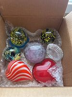 Seven glass paperweights in a box with bubble wrap showing various designs and shapes including floral and heart-shaped.