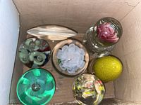 Top view inside a cardboard box showing seven assorted glass paperweights including floral designs and colored pieces.