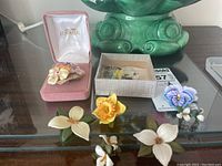 Porcelain brooch displayed in pink velvet box alongside floral leather earrings and flower pins laid on glass table.