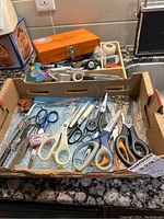 Top-down view of various scissors with handles of different colors, clippers, and small notions in a cardboard box.