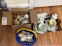 Three boxes and a basket containing assorted decorative shells and plates in various sizes and styles on a hardwood floor.