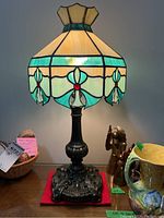 Front view of stained glass lamp lit, showing hexagonal cream, teal, and red pattern and ornate metal base on a red carpet.