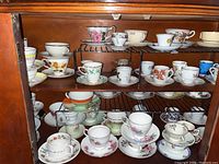 Photo showing wooden cupboard with multiple metal racks holding a variety of vintage porcelain teacups and matching saucers with floral and colored band decoration.