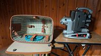 Photo of Bolex M8 projector beside its tweed tone carrying case opened to show checked fabric interior and leather straps.