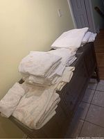 Folded King size linens and sheets with pastel floral designs and plain white linens stacked on a wooden dresser.