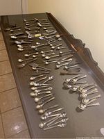 Photo showing a large number of collectible spoons and forks laid out on a wooden table, showcasing variety in handle designs and sizes.