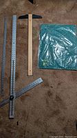 Photo showing the adjustable protracting ruler (aluminum adjustable square), stainless steel yardstick, wooden drafting ruler, and green cutting mat on carpet floor.
