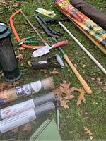 Garden tools and clutter spread on grass including a pruning saw, hand pruners, a hand trowel, small hoes, rolls of garden ground cover fabric, and a soil spreader.