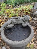 Concrete birdbath with resting cherub figure on rim, showing full top view with basin and surrounding garden