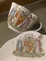 Photo showing the exterior side of the cup and top view of the saucer displaying the colorful royal coat of arms and allegorical figures with 'Coronation 1911' text.