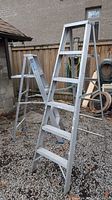 Two aluminum step ladders shown side by side outdoors on gravel ground, near a fence.
