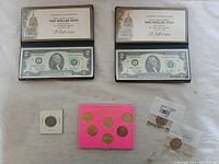 Two $2 bills in protective cases displayed alongside assorted coins including a 1964 Kennedy half dollar, five Lincoln pennies in a pink holder, and two other individual coins.