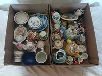Full overview of two cardboard boxes packed with assorted ceramic knick knacks, including plates, cups, small teapots, and figurines.