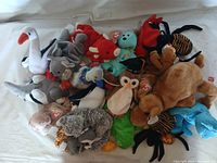 Photo showing a variety of Beanie Babies including a white swan, gray koala, blue bird, brown camel, and others all with Ty tags attached, collected together on a white surface.