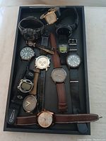 Top view of 10 assorted wristwatches in black tray showing varied bands and watch faces