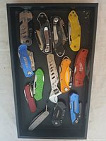 Top-down photo of collection of folding knives and multi-tools arranged in a tray against a white background.
