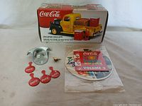Lot contents showing the 1947 Coca-Cola metal pickup truck bank in original box, four red Coca-Cola badges, metal bottle opener, brass emblem, and Coca-Cola music CD in packaging.