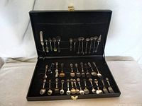 Open black display case showing 36 collector spoons with various designs, arranged in rows secured by elastic bands.