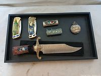 Display tray with all items arranged: fixed blade knife, two metal clips with wildlife paintings, two decorative metal sleeves with stone insets, and a silver pocket watch.