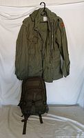 Front view of the olive green military jacket with hood hung on a hanger, and the olive green tactical backpack placed below it.
